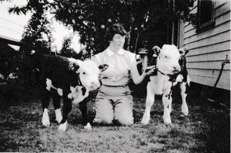 Black and white image of a young woman kneeling between two hereford calves. 