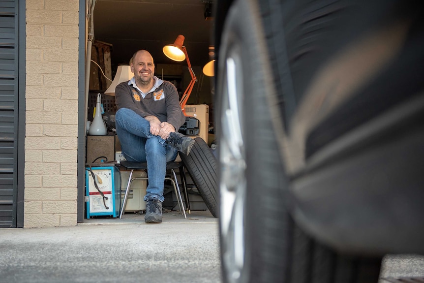 A man sits on a chair in a garage, surrounded by lights. The image is partially impaired by a car.