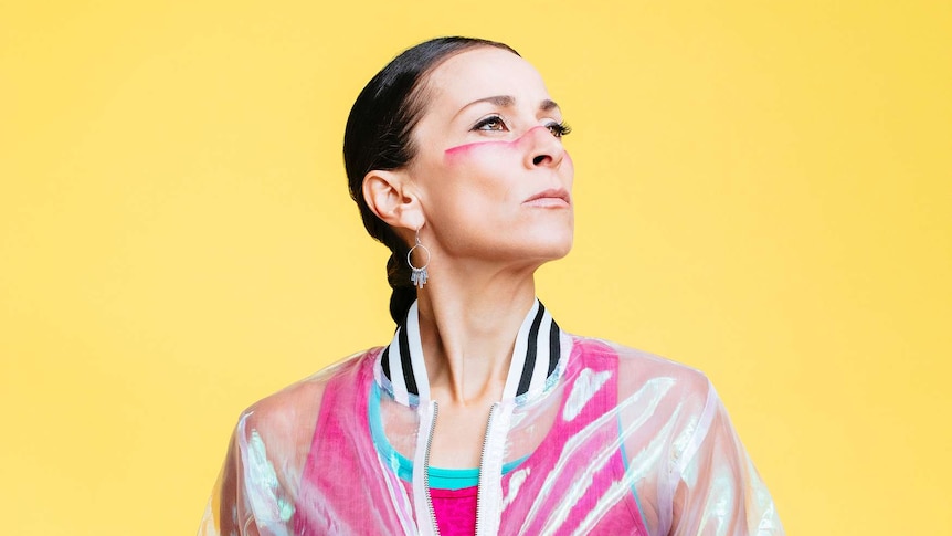 A woman with a pink line of paint across her face stands in front of a yellow backdrop.