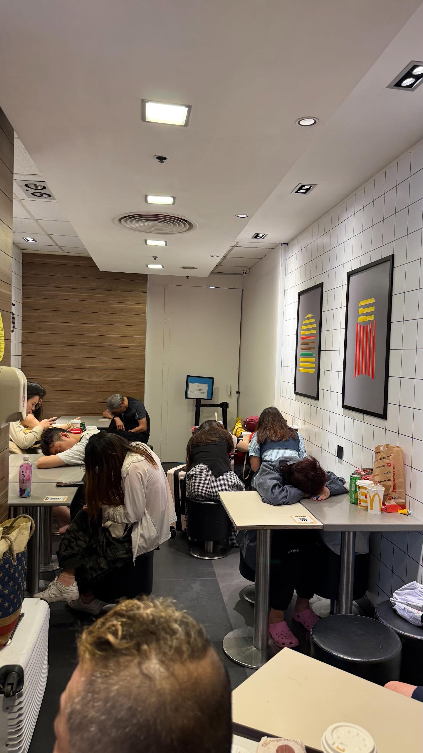 A group of tourists sleep with their heads on small tables inside a well-lit McDonald's outlet.