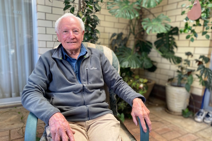 A smiling older man with short, grey hair, sitting in a chair outside.