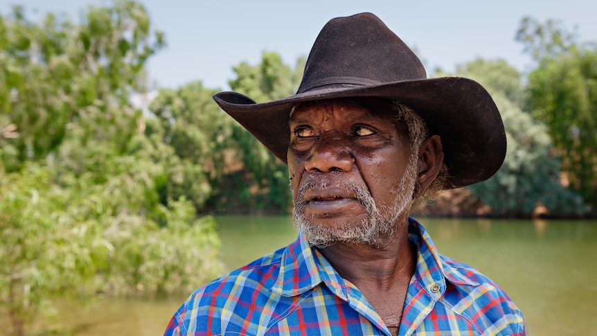 A serious-looking man in an Akubra and flannel shirt looking to the side, with a river and greenery in the background.
