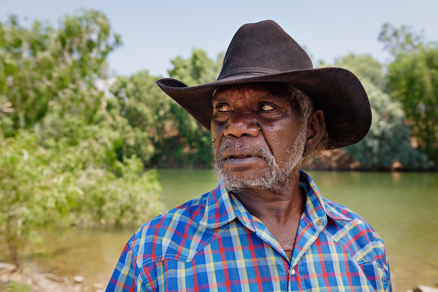 A serious-looking man in an Akubra and flannel shirt looking to the side, with a river and greenery in the background.