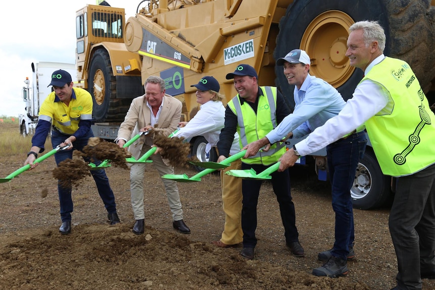 A group of people outside with green shovels digging and smiling at the camera in front of large machinery 