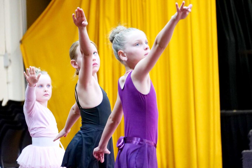 Three young girls stretching their arm toward the sky in ballet class.