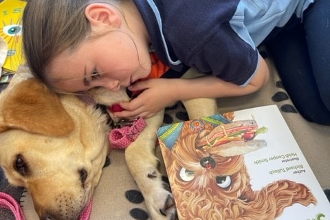 A young school girl lays her head on the side of a large dog, laying on the ground, with a book in front of them.