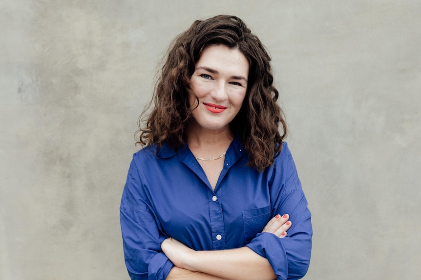 Bridie Jabour, with shoulder-length brown wavy hair, red lipstick, blue shirt and large smile, stands with arms folded.