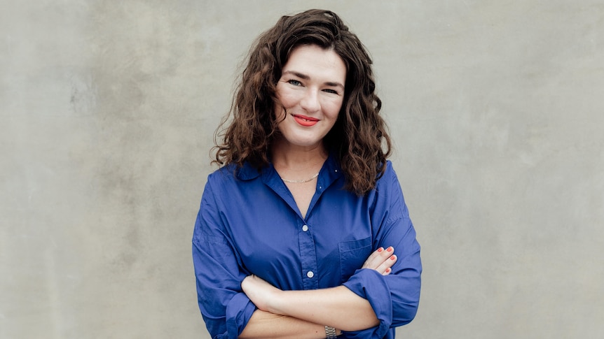 Bridie Jabour, with shoulder-length brown wavy hair, red lipstick, blue shirt and large smile, stands with arms folded.