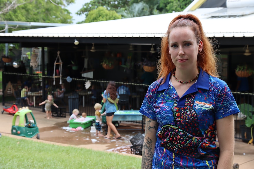 A woman in a blue polo shirt looks seriously at the camera. Behind her children play happily.