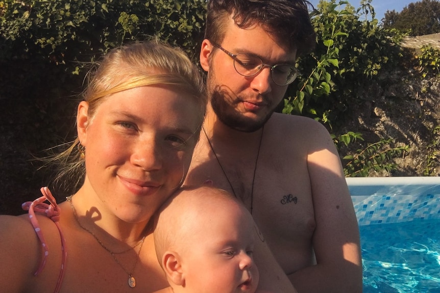 A young woman and man holding an infant near a swimming pool.