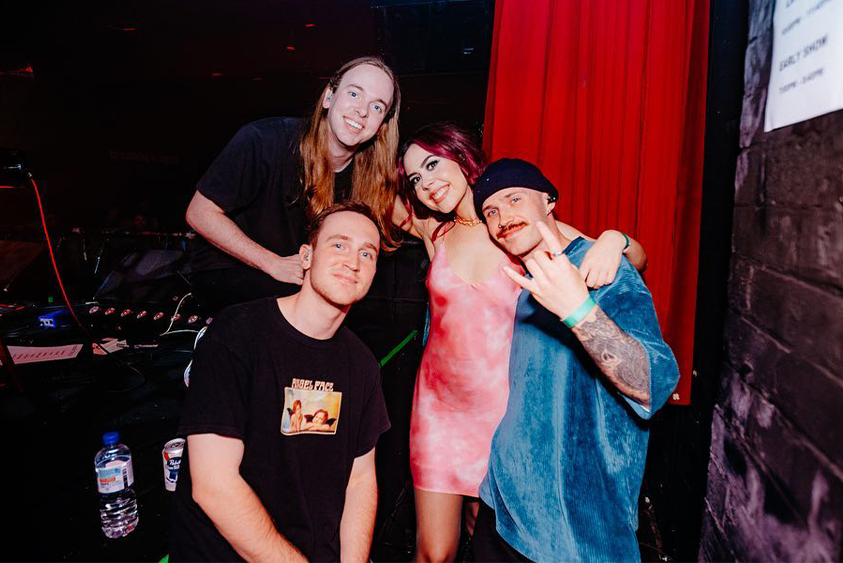 Three men, all wearing t-shirts, and one woman, wearing a pink slip dress, stand backstage