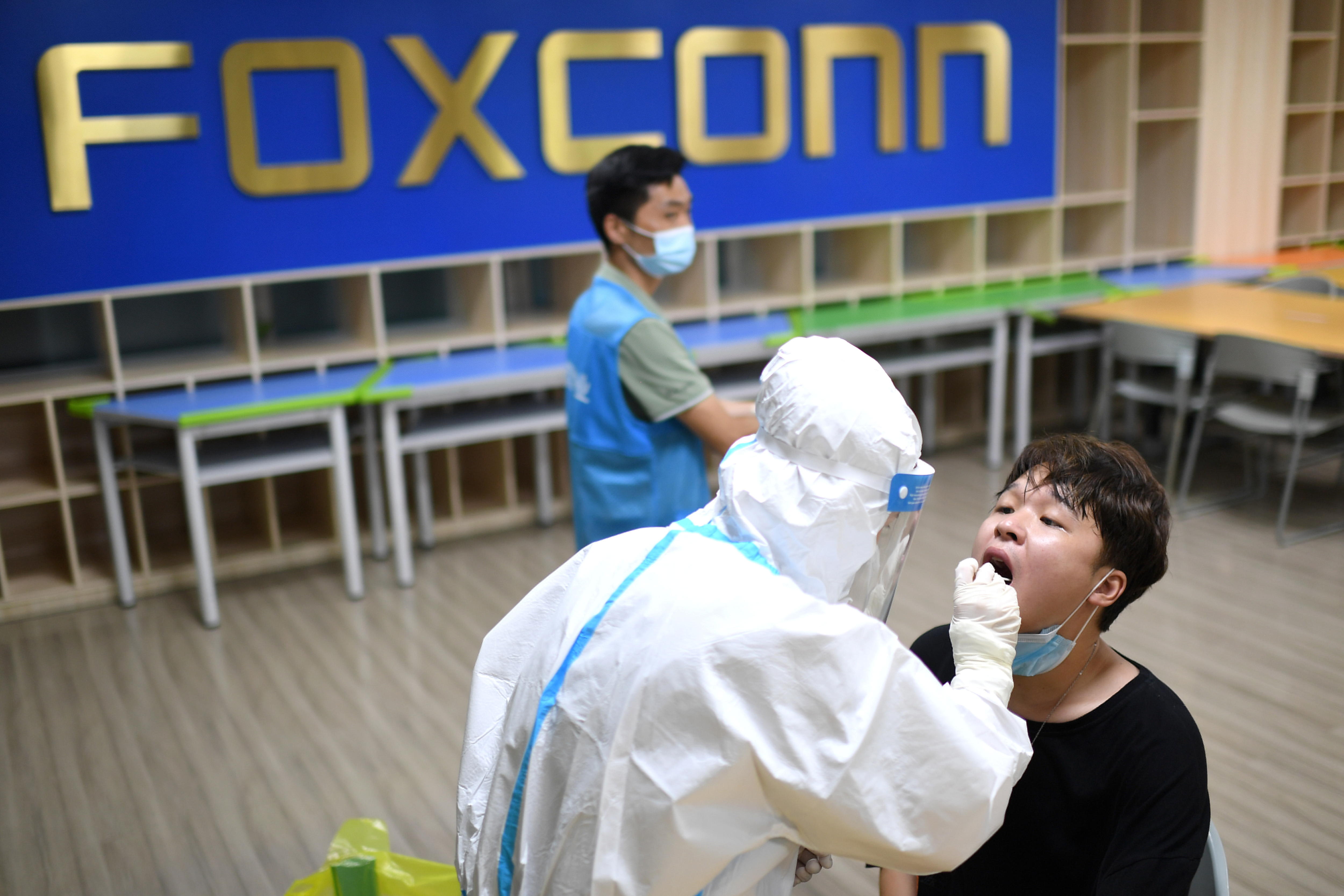 A man sits in a chair and opens his mouth as a person wearing white protective gear does a covid test. 