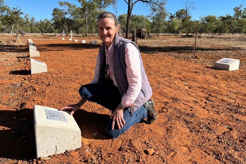 A lady dressed in a blue vest, pink shirt and jeans leans at grave on red soil in outback Queensland.