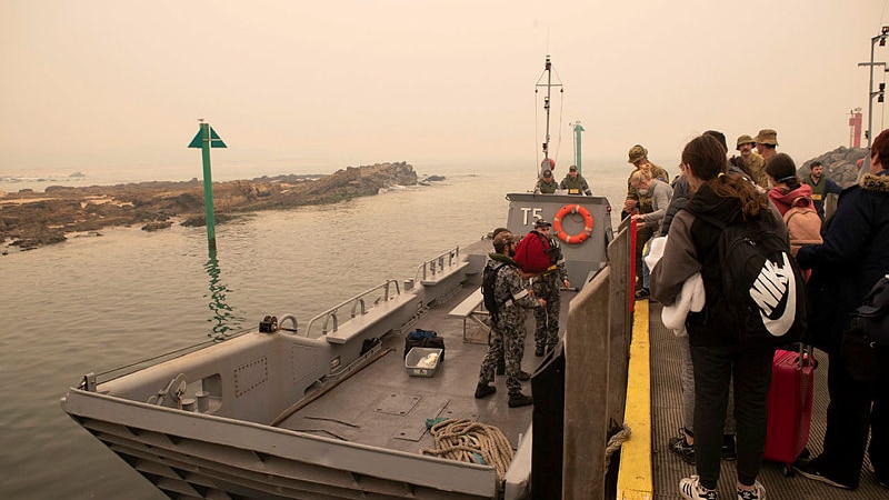About 20 people are seen preparing to board a grey landing craft marked T5. Navy personnel help them aboard.