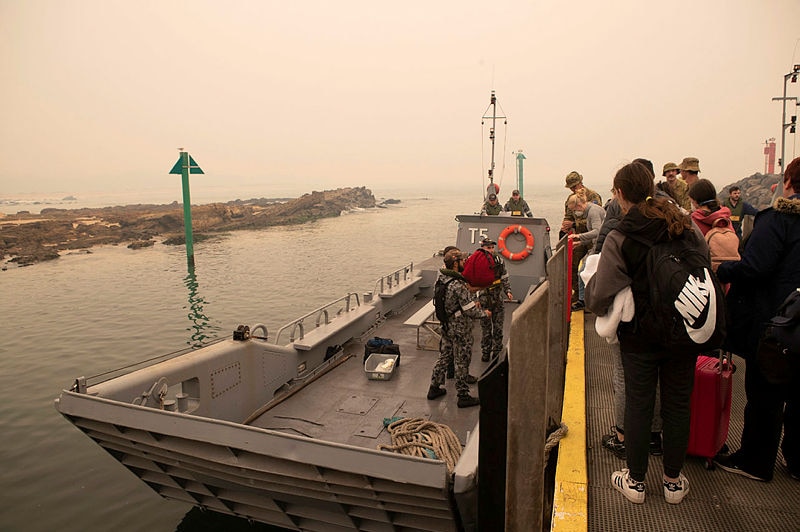 About 20 people are seen preparing to board a grey landing craft marked T5. Navy personnel help them aboard.