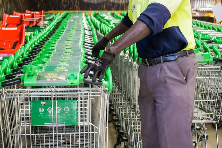 A man wearing hi-vis pushes a line of trolleys into a storage area at a supermarket