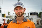 Peter West, wearing an orange overalls and a cap, looks ahead, with boats in the background. 