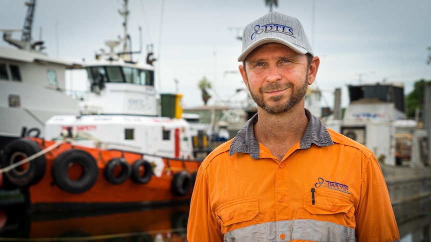 Peter West, wearing an orange overalls and a cap, looks ahead, with boats in the background. 