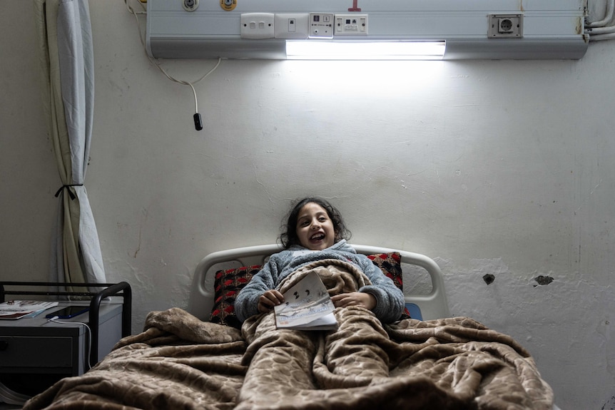 A young girl lays on a hospital bed under a fluroscent hospital light holding a pamphlet.