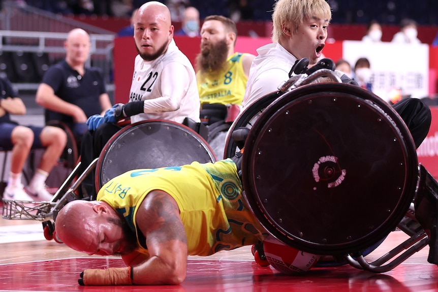 A bald man in a gold singlet falls over in wheelchair.