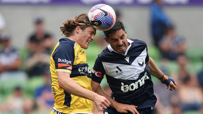Two male soccer players, one wearing yellow and blue and the other wearing navy and white, head the ball during a game
