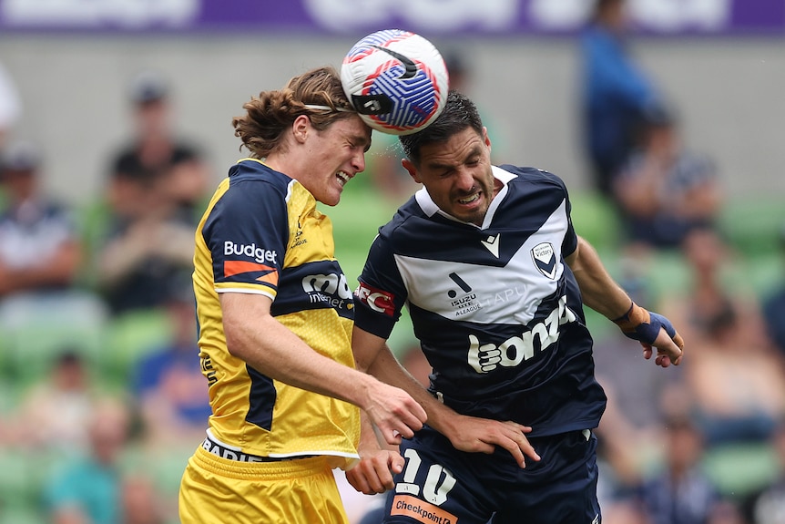 Two male soccer players, one wearing yellow and blue and the other wearing navy and white, head the ball during a game
