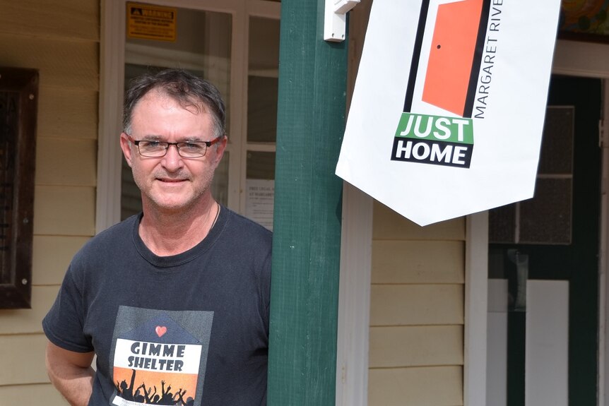Man wearing t-shirt with Gimme Shelter on it stands by a banner for Just Home Margaret River