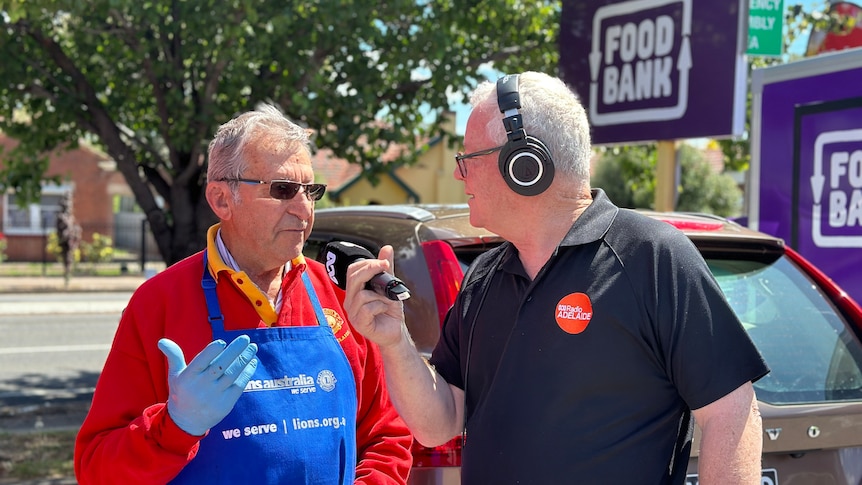man in red shirt and blue apron wearing sunglasses talks to grey haire dman in black polo with microphone