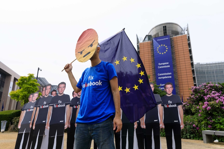 A man with an angry emoji mask stands in front of cardboard cut outs of Mark Zuckerberg.