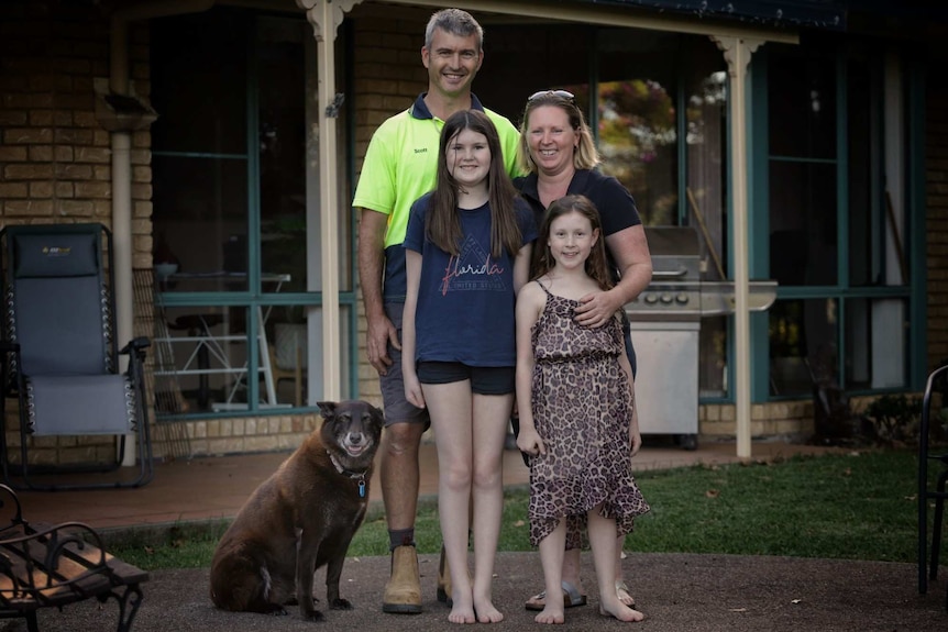 Kristy and Scott Aitken and their two daughters and dog smiles as they stand in front of their home.