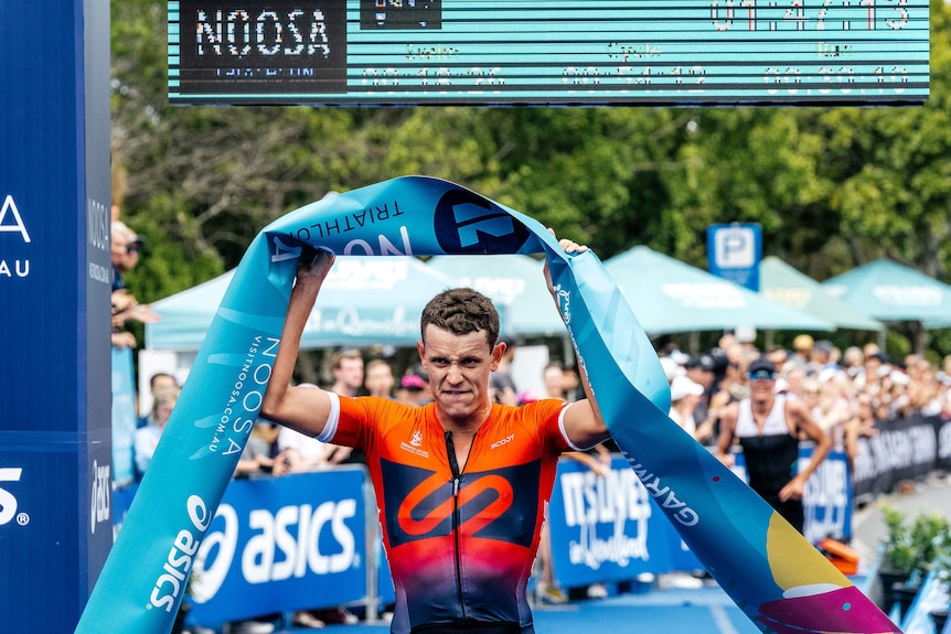 Luke Willian holds a blue banner above his head and grimaces wearing a red triathlon suit