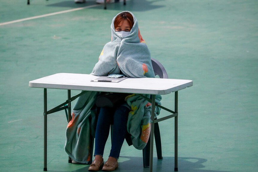 A woman in a face mask wrapped in a blanket sits at a desk in a courtyard