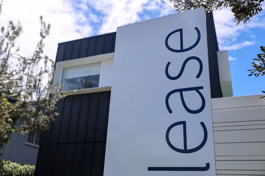 A big white sign that reads "lease" in front of a dark-coloured house.