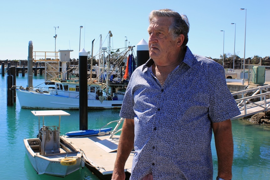 A man with grey hair, wearing a blue shirt, standing at a marina.