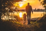 Shot from behind of man walking with child as sun sets in background.