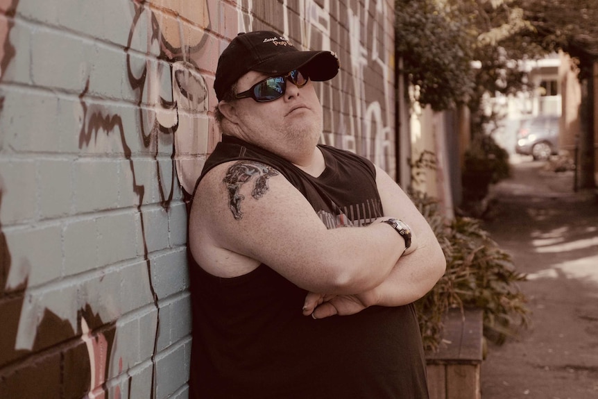 A man in a black cap and sunglasses poses in an alley way