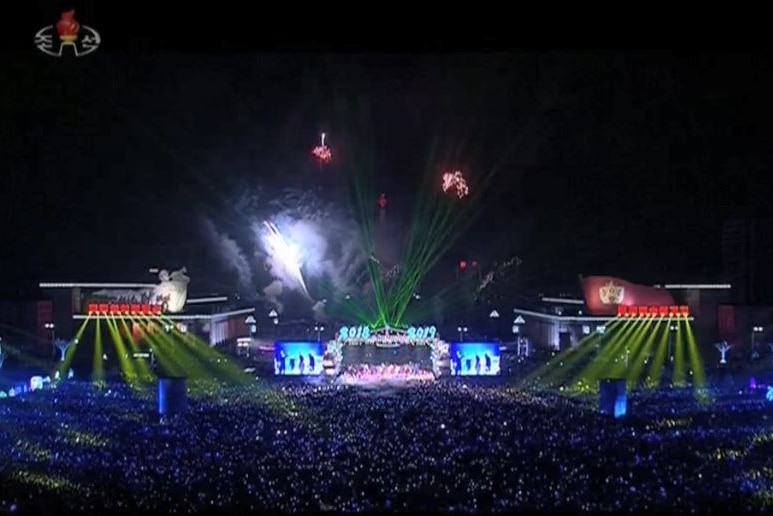 Tens of thousands of people gather before a large stage with performers as fireworks and green lasers fire from the stage's roof