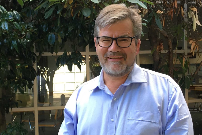 A bespectacled man with greying hair and beard standing in a leafy setting.