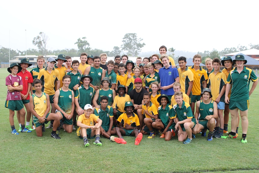 A group of young boys in green and gold rugby uniforms gather around a man with a red cap in a black shirt