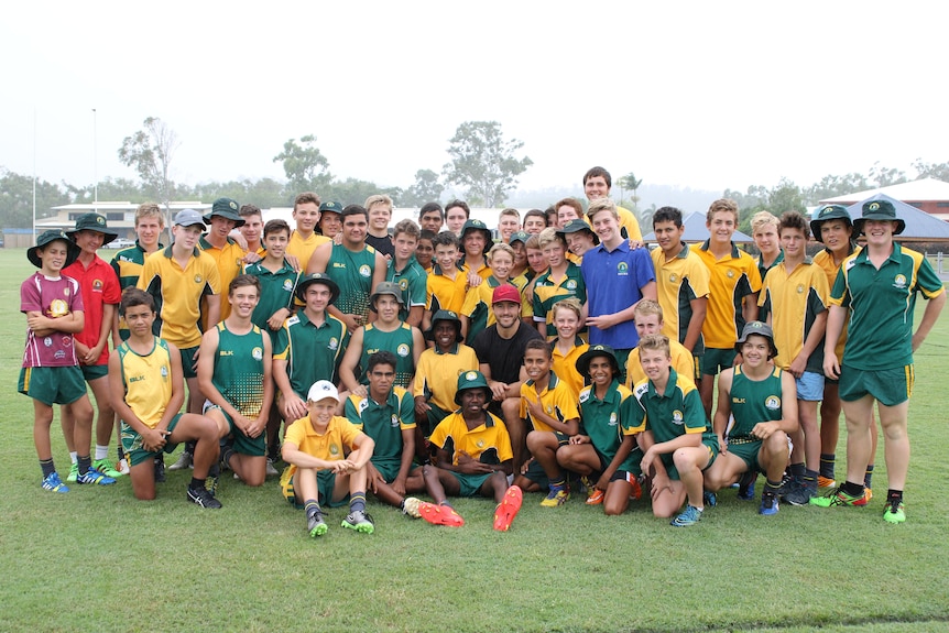 Un groupe de jeunes garçons en uniforme de rugby vert et or se rassemblent autour d'un homme avec une casquette rouge dans une chemise noire