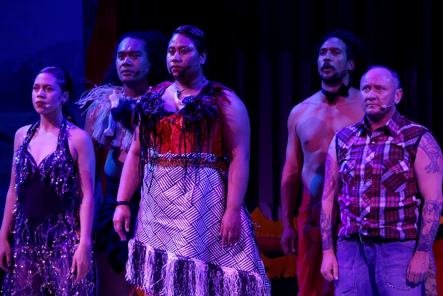 Purple-lit darkened stage with five performers standing front of stage but off camera, in group.