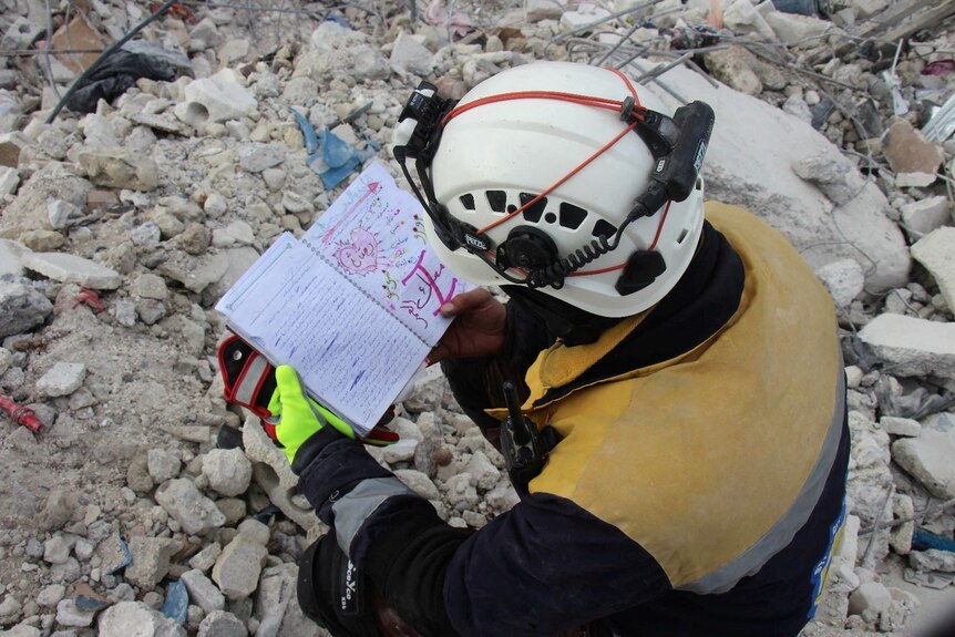 A rescuer sits in rubble reading a notebook he found. 