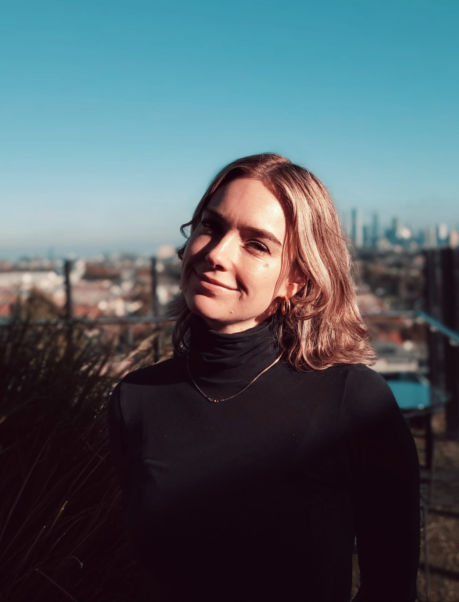 A blonde woman in dark turtleneck poses against a morning city skyline.