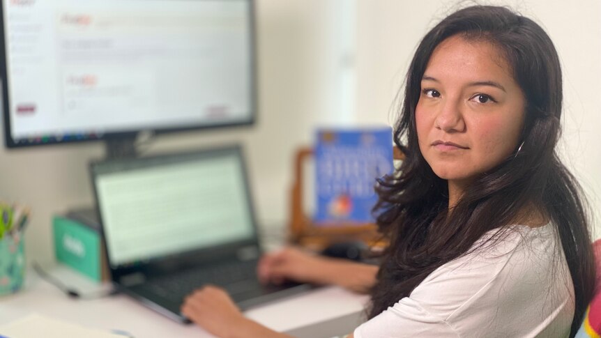 A young woman with long dark hair wearing a white t-shirt sits at a desk in front of a laptop and monitor.