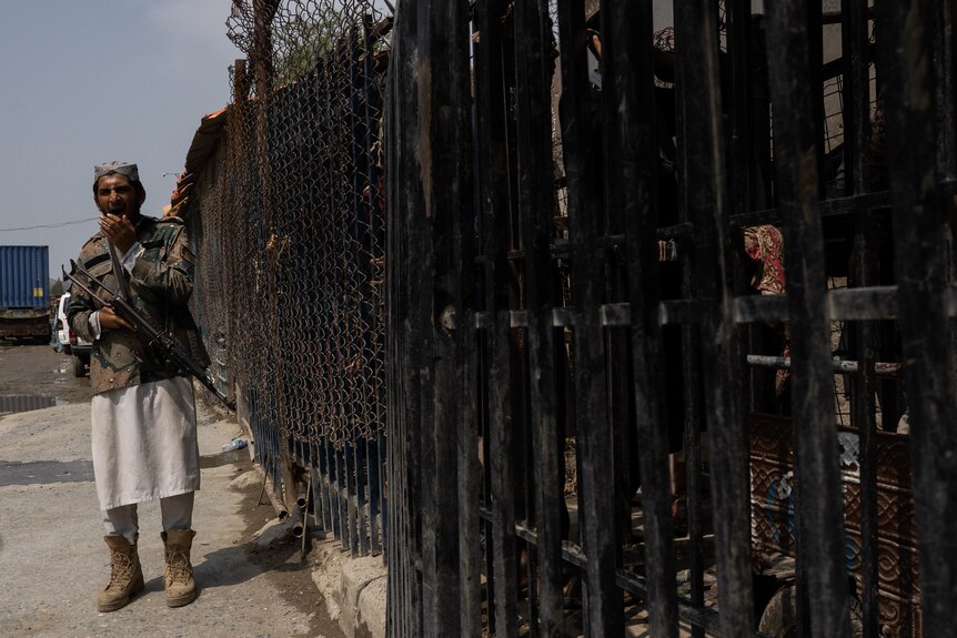 A man holding a huge machine guns stands yawning next to a black fence