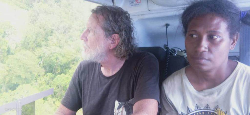 A man and a woman pictured inside a helicopter, both looking out the door at the green forest below