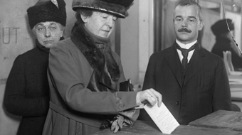 A woman with a large hat dressed in early 20th century fashion looks down as she puts her ballot in a box.