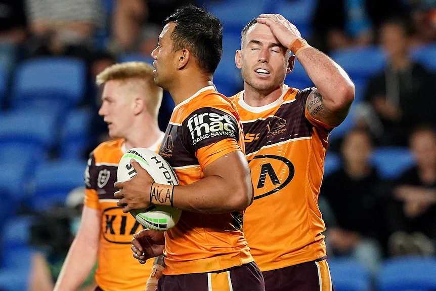 A Brisbane Broncos NRL player puts his hand on his head and closes his eyes as he stands next to a teammate holding the ball.