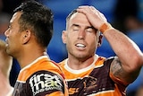 A Brisbane Broncos NRL player puts his hand on his head and closes his eyes as he stands next to a teammate holding the ball.