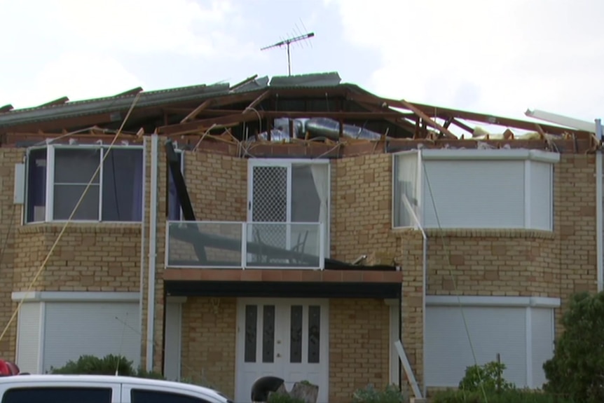 A two-storey house with a damaged roof sitsd in the foreground with grey skies overhead.